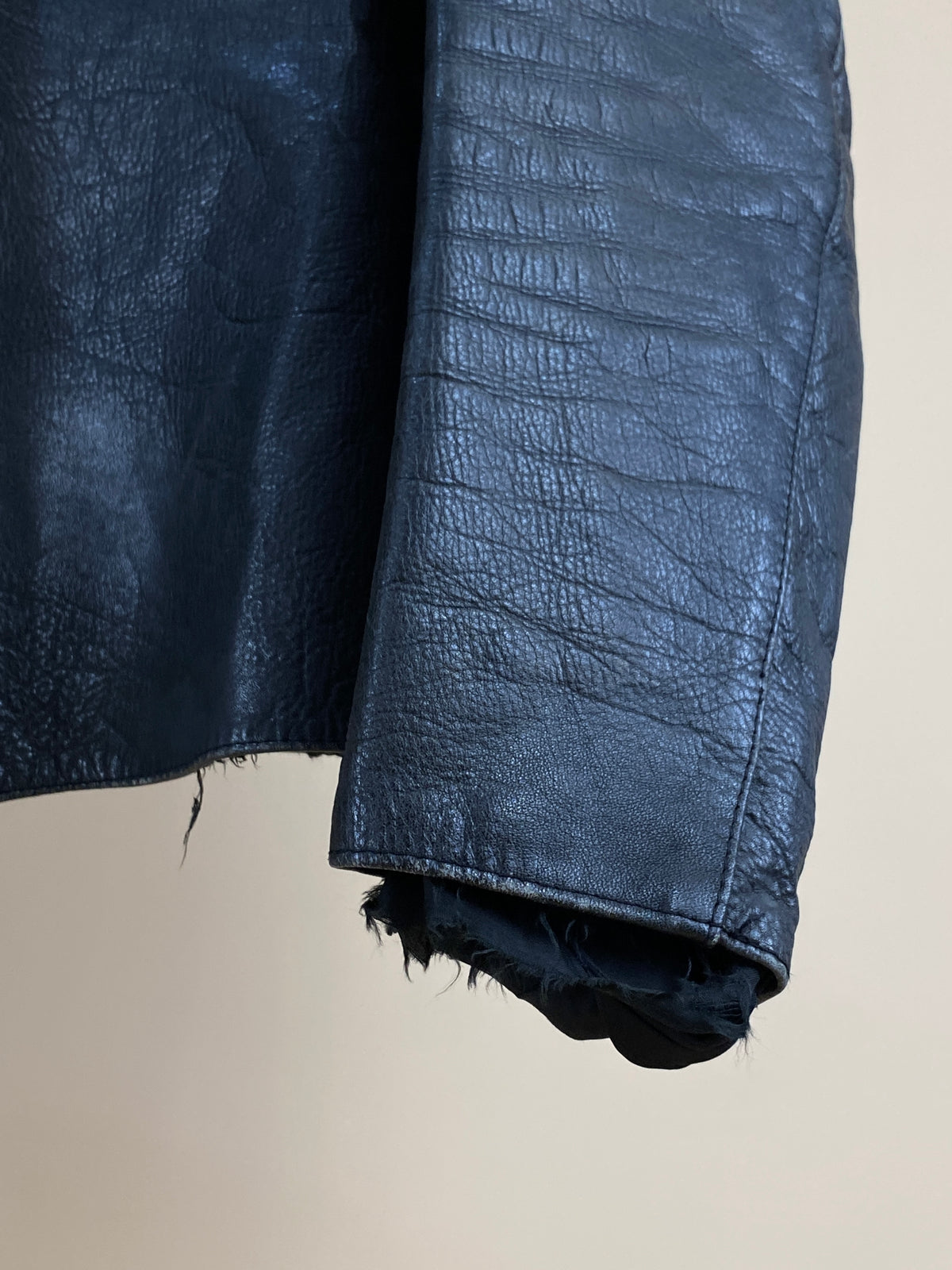 A close-up of the sleeve and hem of the Arkive Vintage 1940s Black Leather Cafe Racer Jacket (size S) showcases its textured surface and frayed edges against a plain beige background.