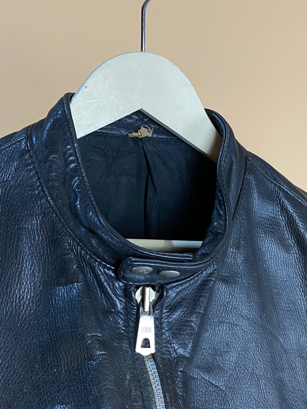 Close-up of the 1940s Black Leather Cafe Racer Jacket - S by Arkive Vintage hanging on a hanger, highlighting the top section with a partially closed front zipper and textured black leather. Inspired by vintage cafe racer jackets, this piece channels classic mid-20th-century style. The background is plain and neutral in color.