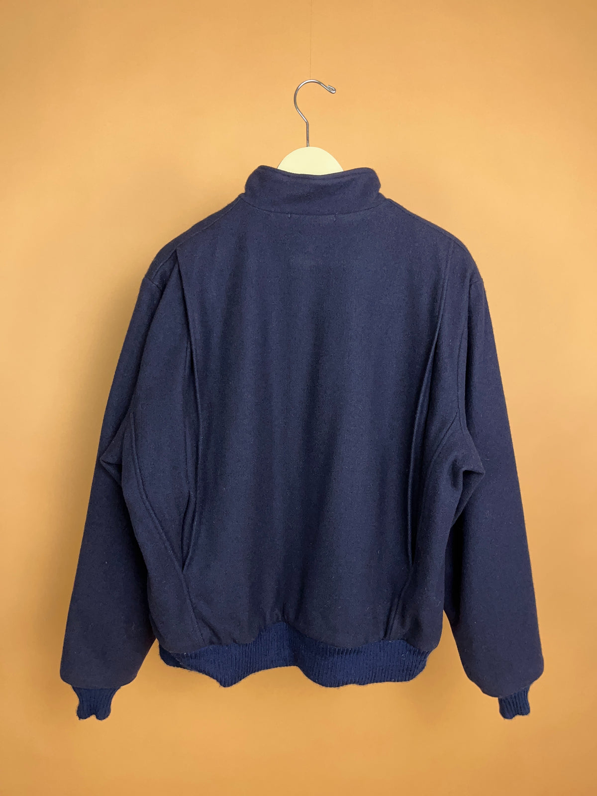 A 1970s Woolrich 5944B navy pure wool bomber jacket with tartan lining, made in the USA and offered by Arkive Vintage, is displayed hanging on a wooden hanger against a plain light brown background, showcasing its back and long sleeves.