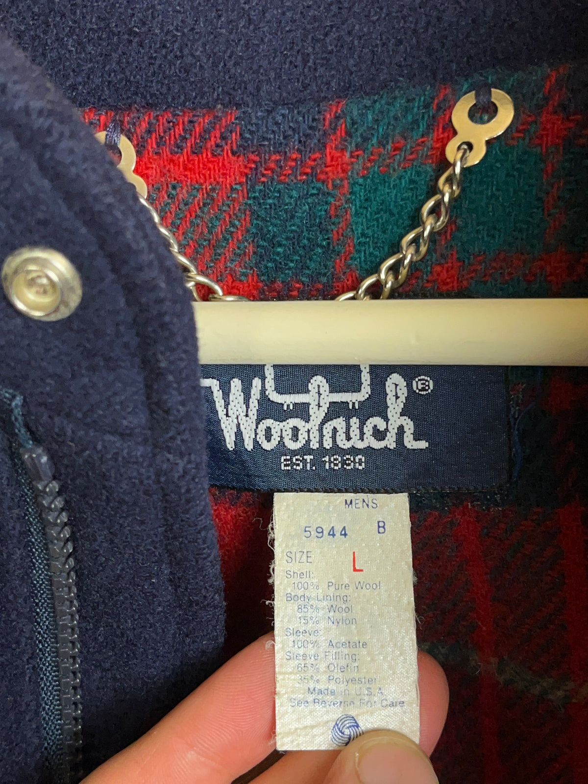 A close-up of the Arkive Vintage 1970s Woolrich 5944B Navy Pure Wool Tartan Lined Bomber Jacket label and size tag, displaying “Woolrich Est. 1830” along with a smaller tag listing materials and size M. The navy fabric is paired with a classic red and green tartan lining, emphasizing this vintage Made in USA wool bomber jacket’s timeless style.