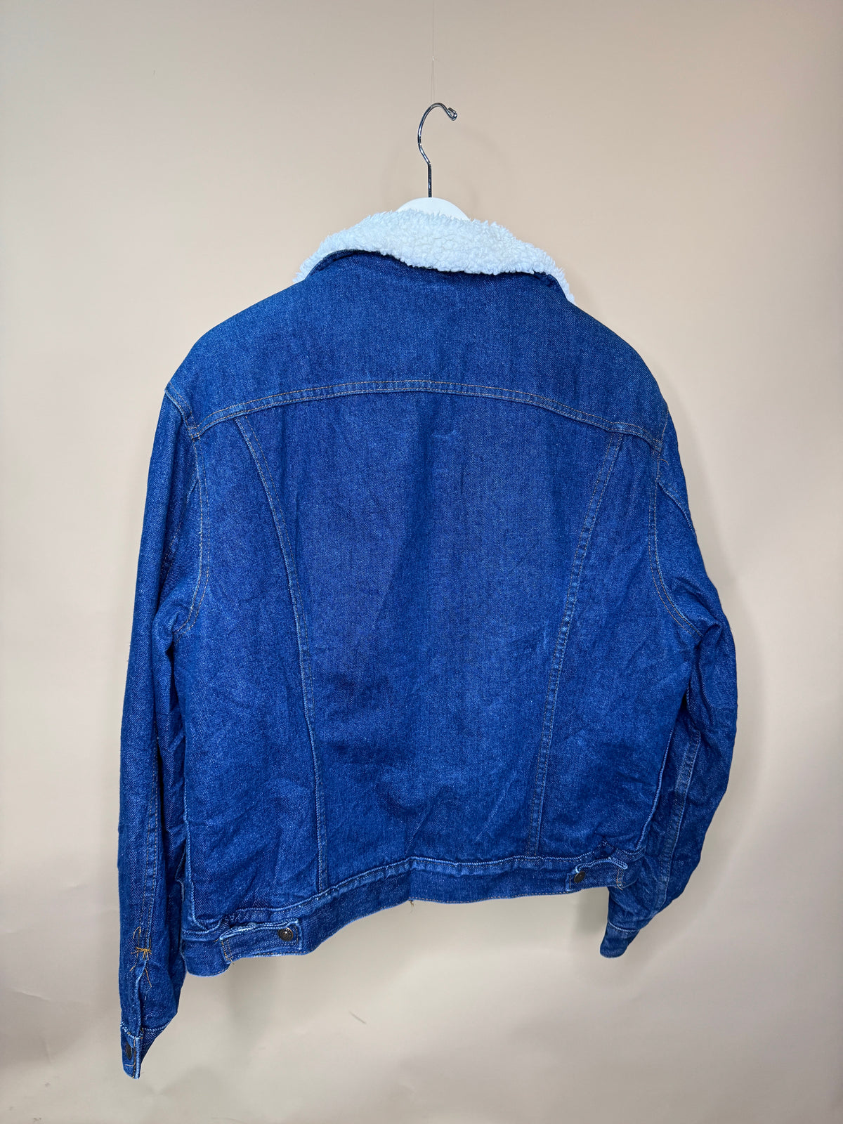 A 1980s Levi’s Boa Lined Stone Wash Denim Jacket by Arkive Vintage, crafted from 100% cotton denim with a white sherpa collar, in Size M/L, hangs on a hanger against a plain beige background, shown from the back.