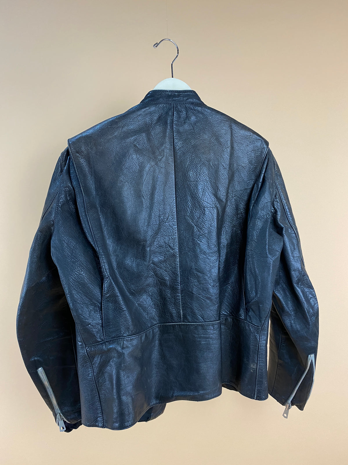 The Arkive Vintage 1940s Black Leather Cafe Racer Jacket - S is displayed from the back, hanging on a white hanger against a beige wall. This classic piece features visible sleeve zippers and a signature Nauru collar.
