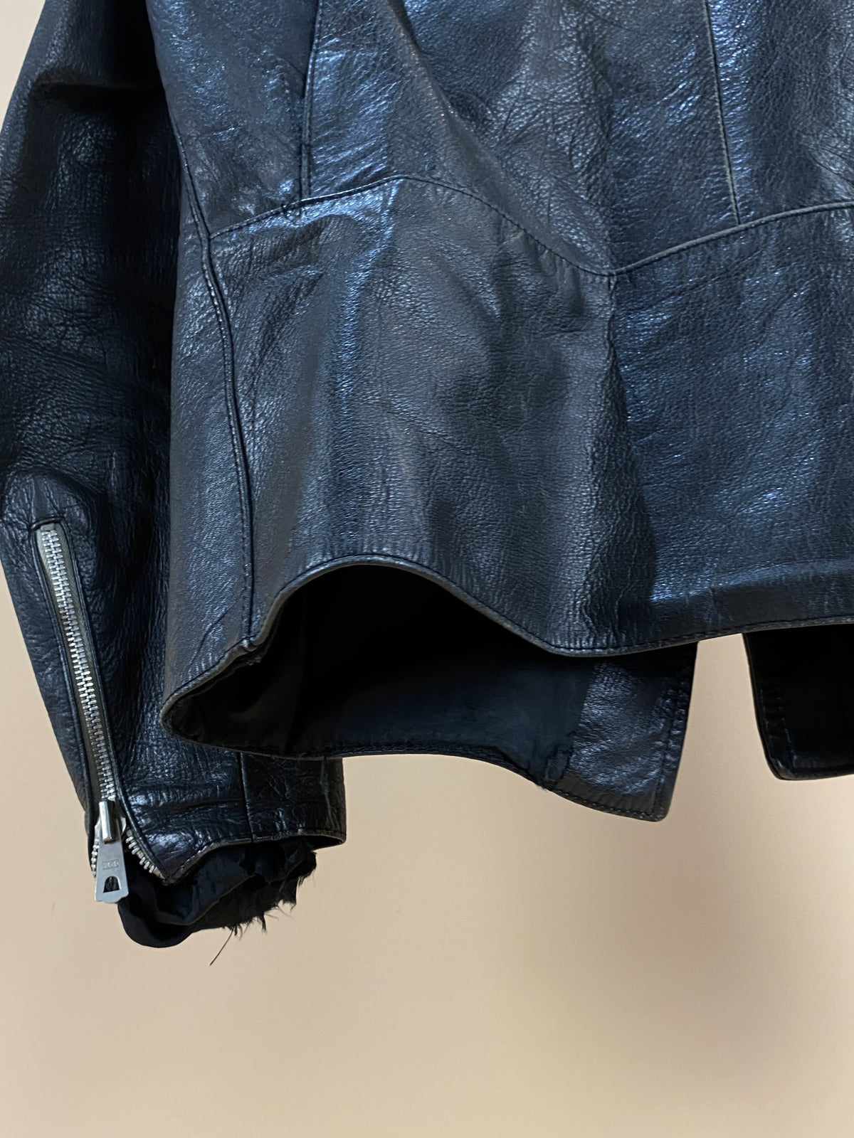 A close-up of the Arkive Vintage 1940s Black Leather Cafe Racer Jacket (size S) hanging, highlighting its textured leather, a side zipper on the sleeve, and a glimpse of the unfinished inner lining.