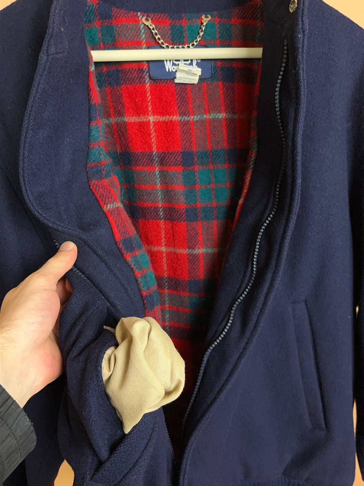 A person holds open the Arkive Vintage 1970s Woolrich 5944B Navy Pure Wool Tartan Lined Bomber Jacket (Made in USA, size M), revealing a beige rolled-up cloth in the pocket.