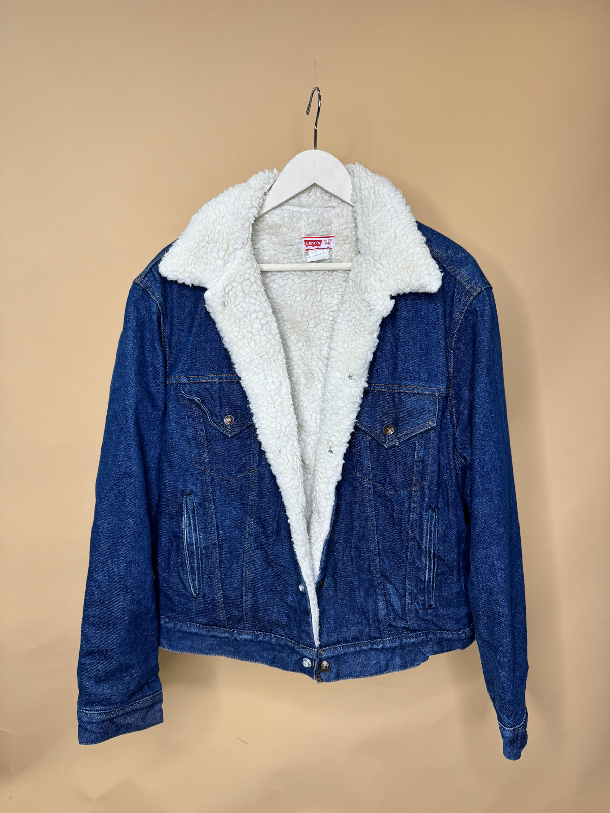 A 1980s Levi’s Boa Lined Stone Wash Denim Jacket from Arkive Vintage, made in the USA from 100% cotton, hangs on a white hanger against a plain tan background. This blue denim jacket features a white sherpa lining, two buttoned chest pockets, and visible stitching details. Available in size M/L (46).